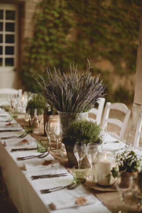 a burlap wedding table runner with some potted greenery as sustainable wedding decor, lavender in a vase and some candles