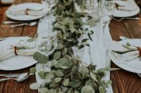 a carefree backyard wedding table setting with neutral linens and a lush eucalytus runner plus candles is a lovely idea