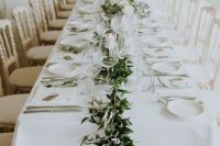 a chic backyard wedding table setting with white linens and plates, with a greenery runner dotted with candles and silver cutlery