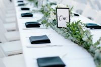 a chic modern wedding table setting with a white tablecloth, navy napkins, a greenery table runner with white blooms