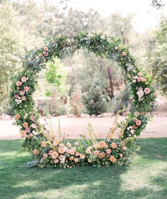 a circle spring wedding arch decorated with greenery and peachy and pink blooms all around