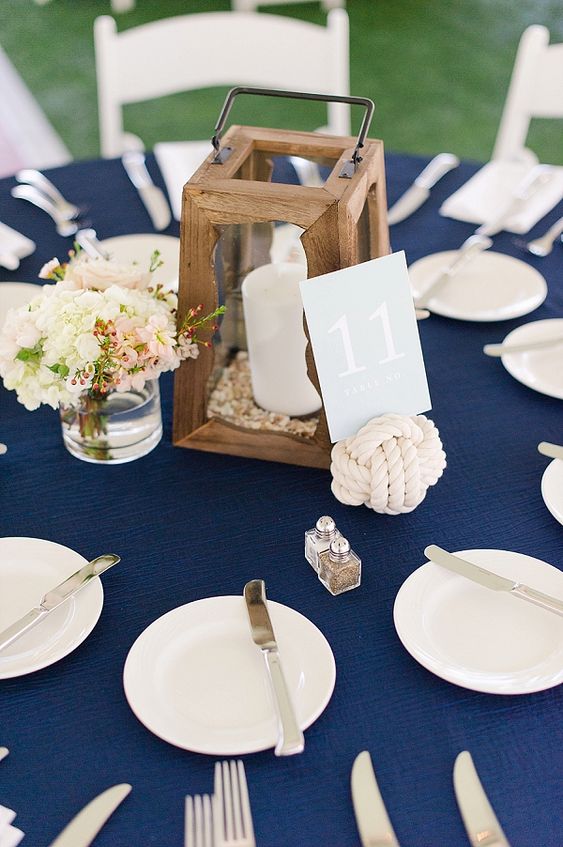 a classic navy tablescape with a navy tablecloth, a white floral centerpiece, a wooden candle lantern and a rope table number stand
