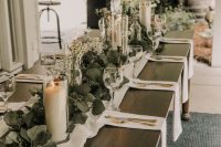 a classic wedding table setting with white linens, a lush greenery runner, candles and daisy centerpieces plus gold cutlery
