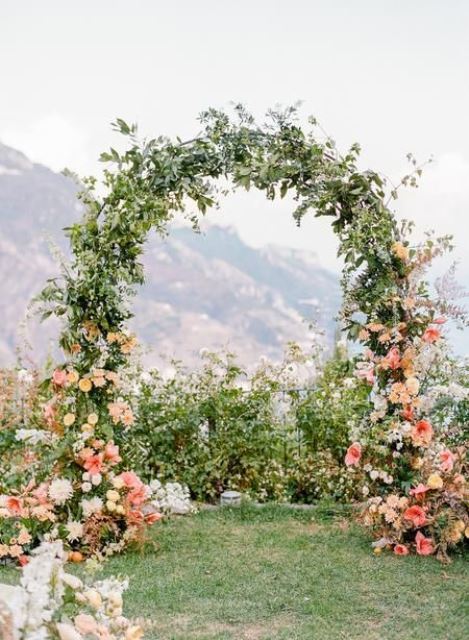 a cool spring wedding arch covered with greenery and decorated with peachy and coral blooms