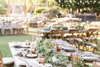 a cozy backyard wedding tablescape with copper mugs and copper candle lanterns, a greenery runner, white plates and napkins plus copper cutlery