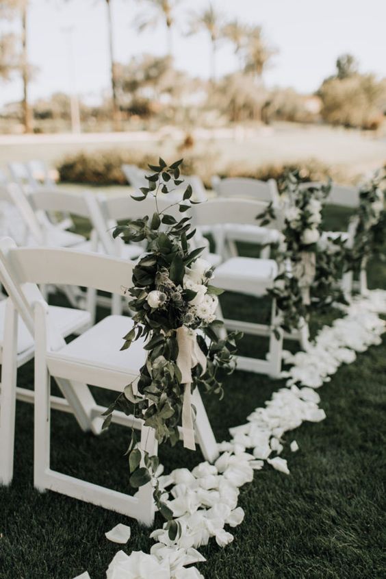 a fresh and edgy spring wedding aisle with white petals on the ground, with white blooms and greenery arrangements on the chairs