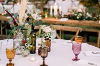 a modern boho backyard wedding tablescape with colorful glasses, pastel blooms with greenery and pampas grass plus candles