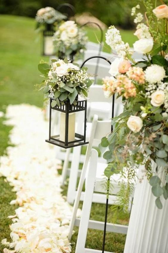 a pretty and chic spring wedding aisle with neutral petals on the ground, with candle lanterns decorated with greenery and white blooms