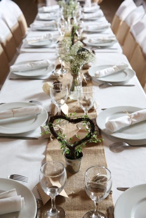 a pretty and cute neutral rustic wedding tablescape with a burlap runner, neutral linens, baby’s breath and a vine heart with greenery