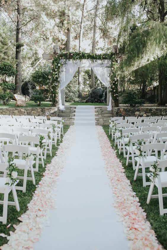 a pretty secret garden wedding aisle with a white liner blush petals white blooms and greenery on the chairs and a matching arch