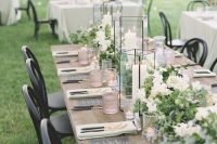 a rustic backyard wedding table setting with an uncovered table, white blooms and greenery, candle lanterns and blush glasses plus black cutlery