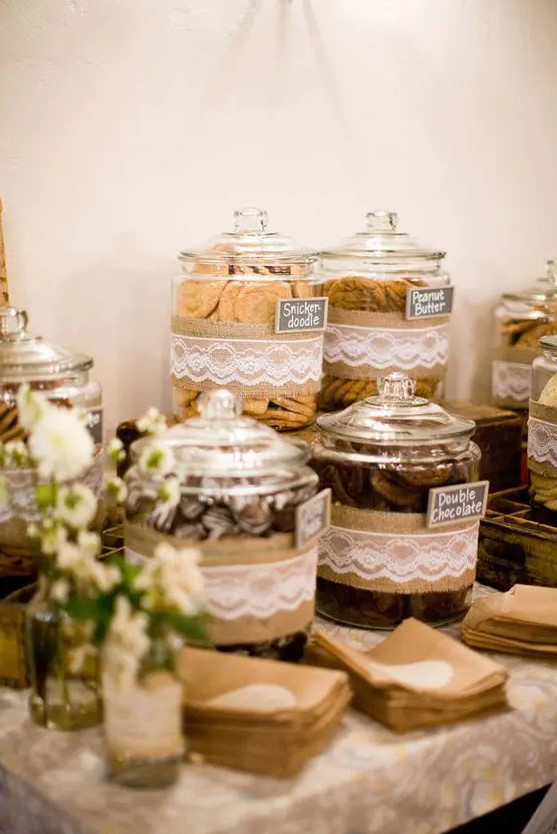 a rustic dessert table with large jars covered in burlap and lace, with neutral blooms and greenery and paper napkins