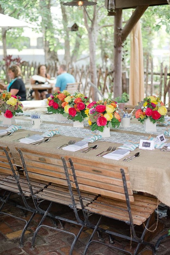 a rustic tablescape covered with burlap, a colorful printed runner, bright blooms in neutral vases and simple napkins