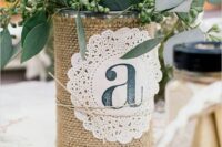 a rustic wedding centerpiece of a jar wrapped with burlap and with a doily, with greenery and blooms is a cool idea for a rustic wedding