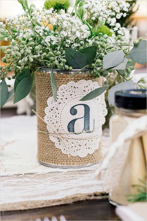 a rustic wedding centerpiece of a jar wrapped with burlap and with a doily, with greenery and blooms is a cool idea for a rustic wedding