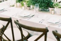 a simple and lovely backyard wedding table setting with potted greenery centerpieces and candles, neutral plates and linens and white chairs