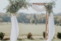 a simple rustic spring wedding arch with flowy white fabric and lots of greenery looks fresh and cool