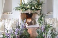 a sophisticated spring wedding aisle with lush greenery and lilac and purple blooms plus a lush floral arrangement on the mantel is jaw-dropping