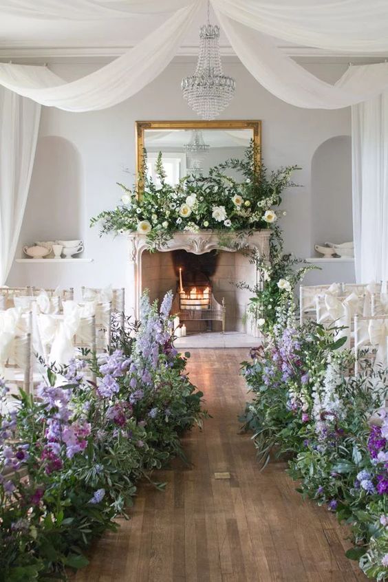 a sophisticated spring wedding aisle with lush greenery and lilac and purple blooms plus a lush floral arrangement on the mantel is jaw-dropping