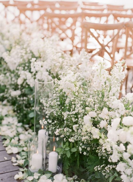 a super bold and lush spring wedding aisle with various greenery and lots of white blooms and glasses with candles is gorgeous