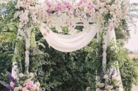 a super lush spring wedding arch decorated with white fabric, white, pink, blue and purple blooms and much greenery