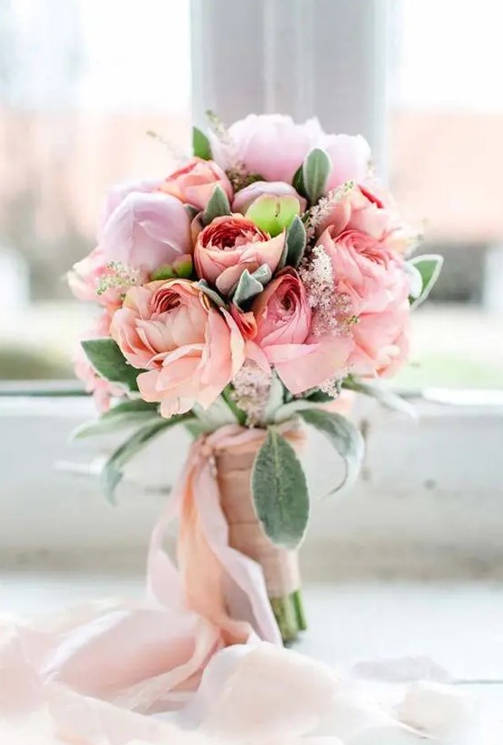a sweet pastel spring wedding bouquet of peachy and light pink blooms and greenery and light pink ribbons