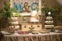 a tiered burlap ruffle tablecloth with wooden stands with cupcakes, greenery, a vintage suticase for displaying a suitcase and monograms over the table