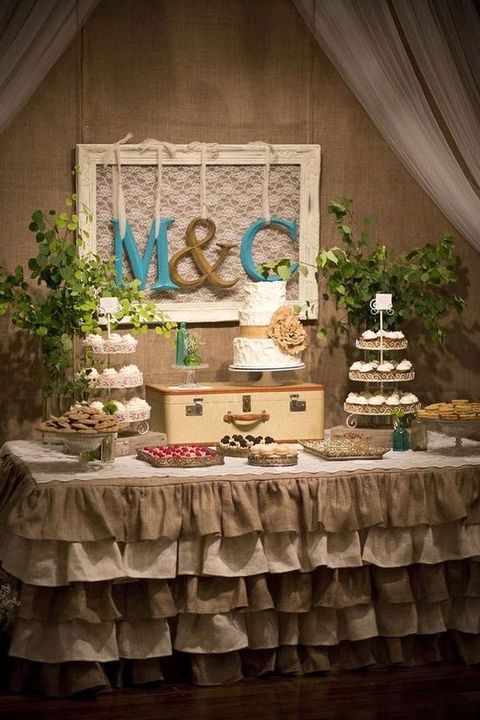 a tiered burlap ruffle tablecloth with wooden stands with cupcakes, greenery, a vintage suticase for displaying a suitcase and monograms over the table