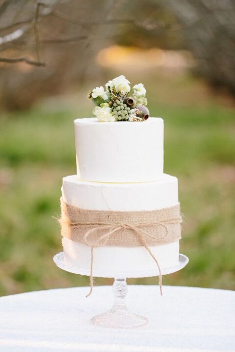 a white wedding cake accented with burlap and twine, with white blooms and seeds pods is a pretty and catchy idea for a rustic wedding