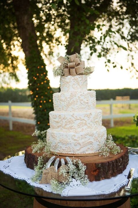 a white wedding cake with beautiful patterns, baby’s breath and a large burlap bow on top for a rustic wedding