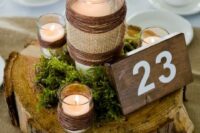 a woodland wedding centerpiece of a wood slice, moss and candles in glasses wrapped with twine and burlap, a table number of wood