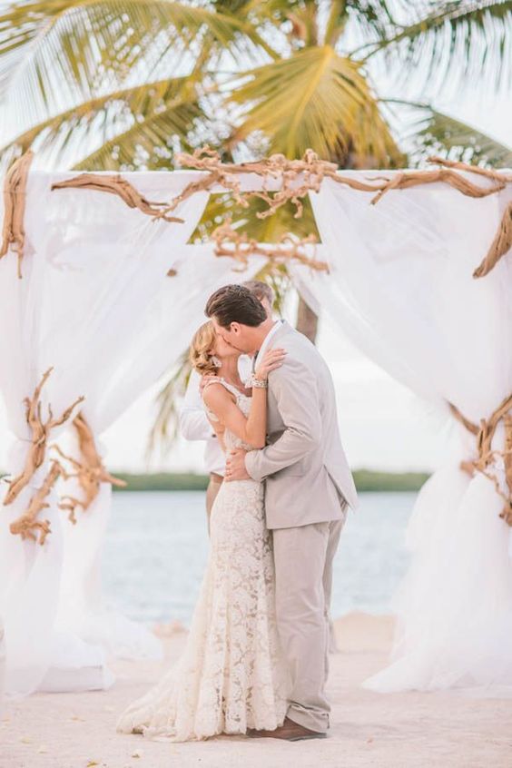 a beach driftwood wedding arch done with white fabric is a very cool idea with a breezy feel