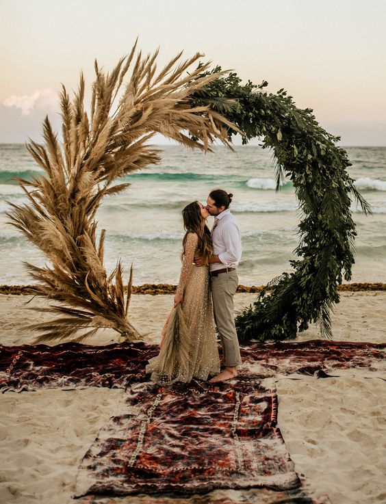 a boho beach circle wedding arch made of greenery and pampas grass plus boho rugs are a super trendy idea for a boho beach wedding
