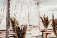 a boho beach wedding space with pampas grass in vases, wooden benches and a wedding arch with dream catchers and macrame