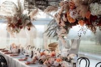a boho chic tablescape done in rust and blush shades with lush florals on the table and over it, wicker lamps, colored napkins and candles