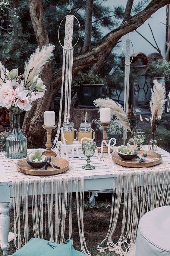 a boho tablescape with a whitewashed table, macrame placemats, colored glasses, candles and florals