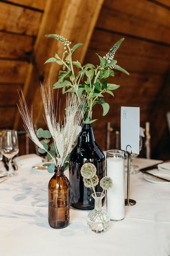 a boho wedding centerpiece of apothecary bottles, a candle, wheat and greenery for a simple look