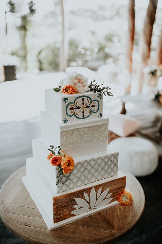 a bright boho wedding cake with various geometric and wood tiers, colorful patterns, bright blooms and greenery