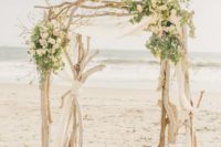 a chic beach driftwood wedding arch with lots of greneery and neutral blooms, white fabric and candle lanterns