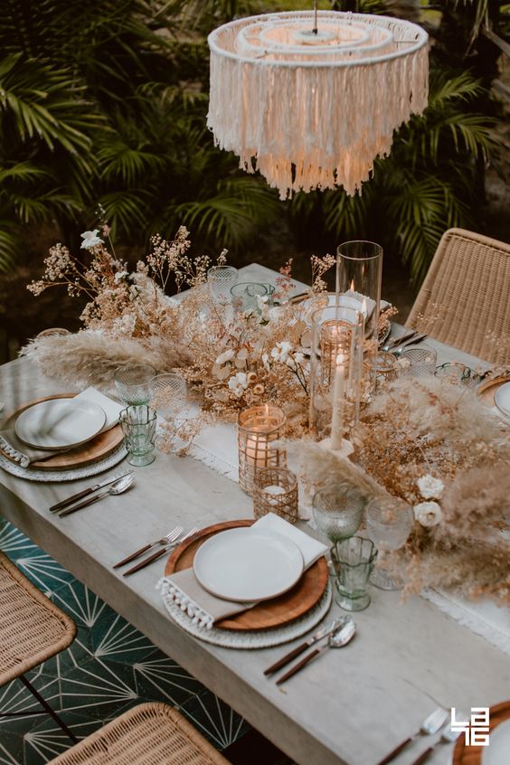 a chic desert boho tablescape with wooden plates, a fringe chandelier, candles, dried herbs and blooms plus pampas grass