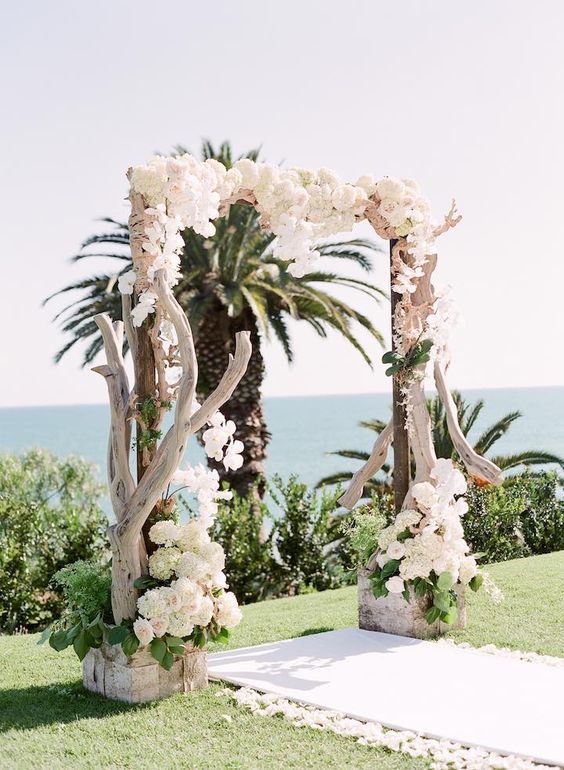 a driftwood wedding arch decorated with white orchids and hydrangeas in a lush way for a refined touch