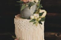 a grey and white wedding cake with gold leaf, macrame detailing, whiet and pink blooms, greenery and a gold topper