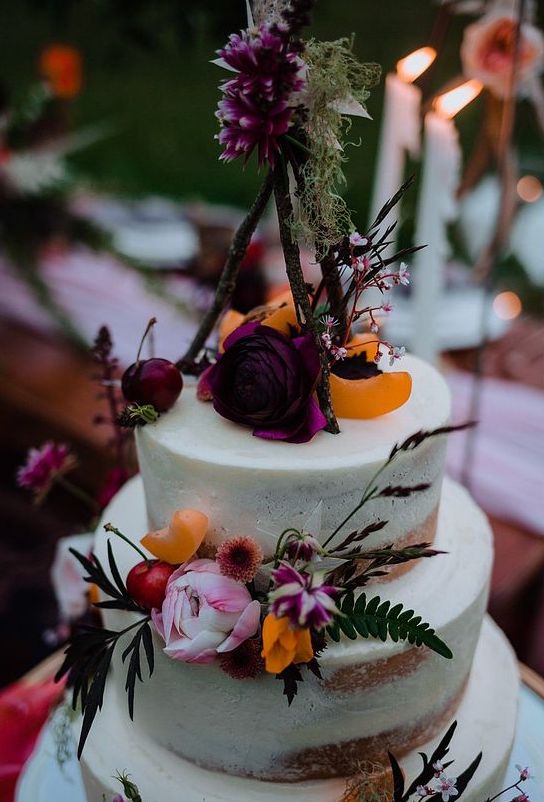 a naked wedding cake with bright blooms, berries, and a teepee on top with moss and bold dramatic flowers