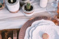 a simple boho chic tablescape with a white runner, patterned plates and chargers, potted cacti, candles and copper cutlery