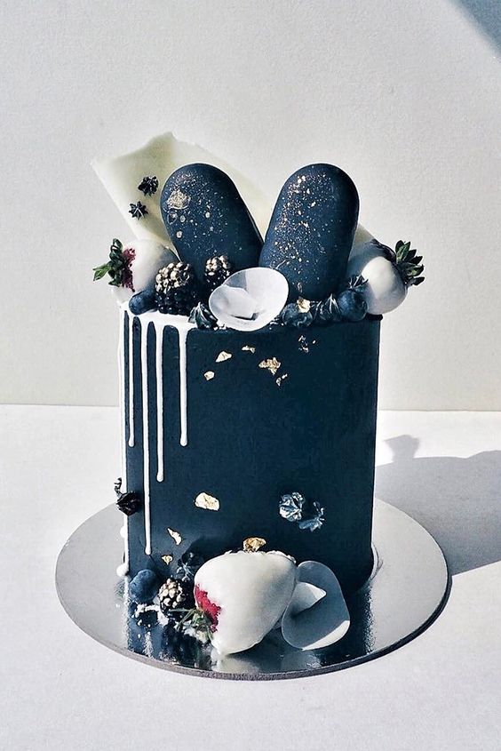 an exquisite black groom's cake with white drip, black and gold popsicles, white chocolate shards, chocolate berries and gold leaf