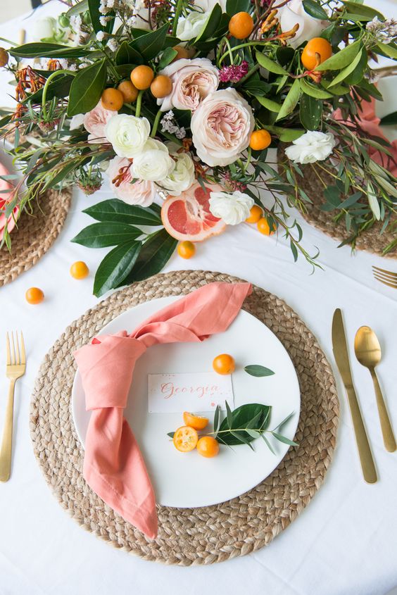 a beautiful spring wedding tablescape with woven placemats, white porcelain, pink napkins, kumquats, a lovely wedding centerpiece of white and blush blooms, greenery and kumquats