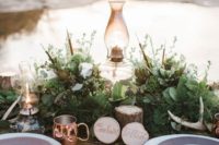a beautiful woodland wedding tablescape with greenery, feathers, white blooms, a lamp, antlers, wood slices and soem feathers