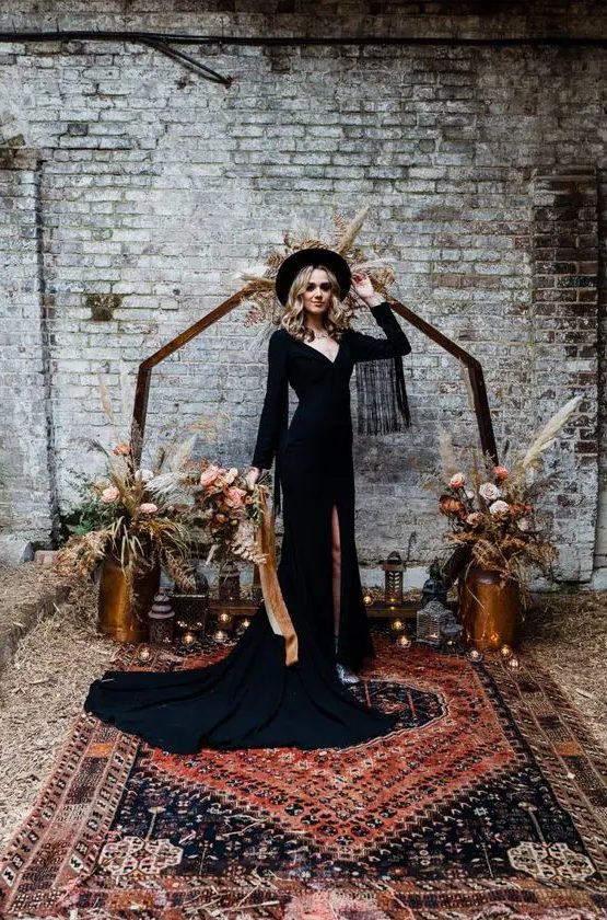 a black A-line boho wedding dress with a deep neckline, fringe sleeves and a train plus a slit, silver boots and a black hat