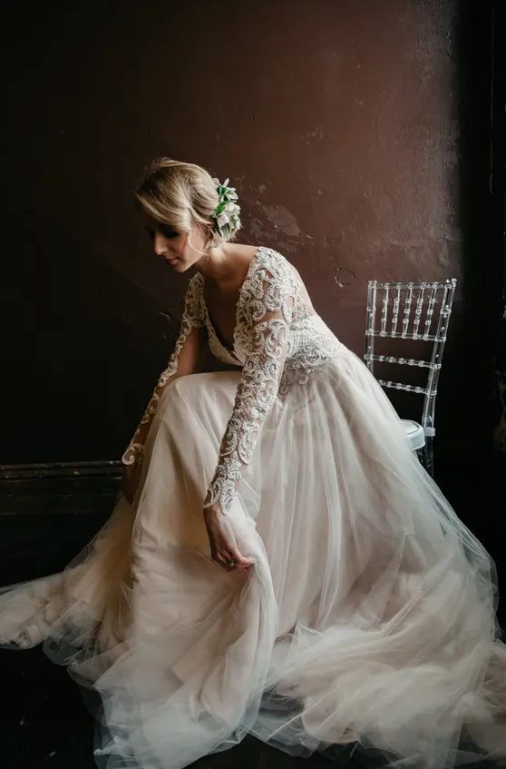 a blush and ivory lace wedding dress with a deep V-neckline and long sleeves and a messy updo with blooms