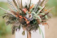 a boho fall wedding bouquet made of feathers, bright blooms, greenery and with ribbons looks unusual and catchy